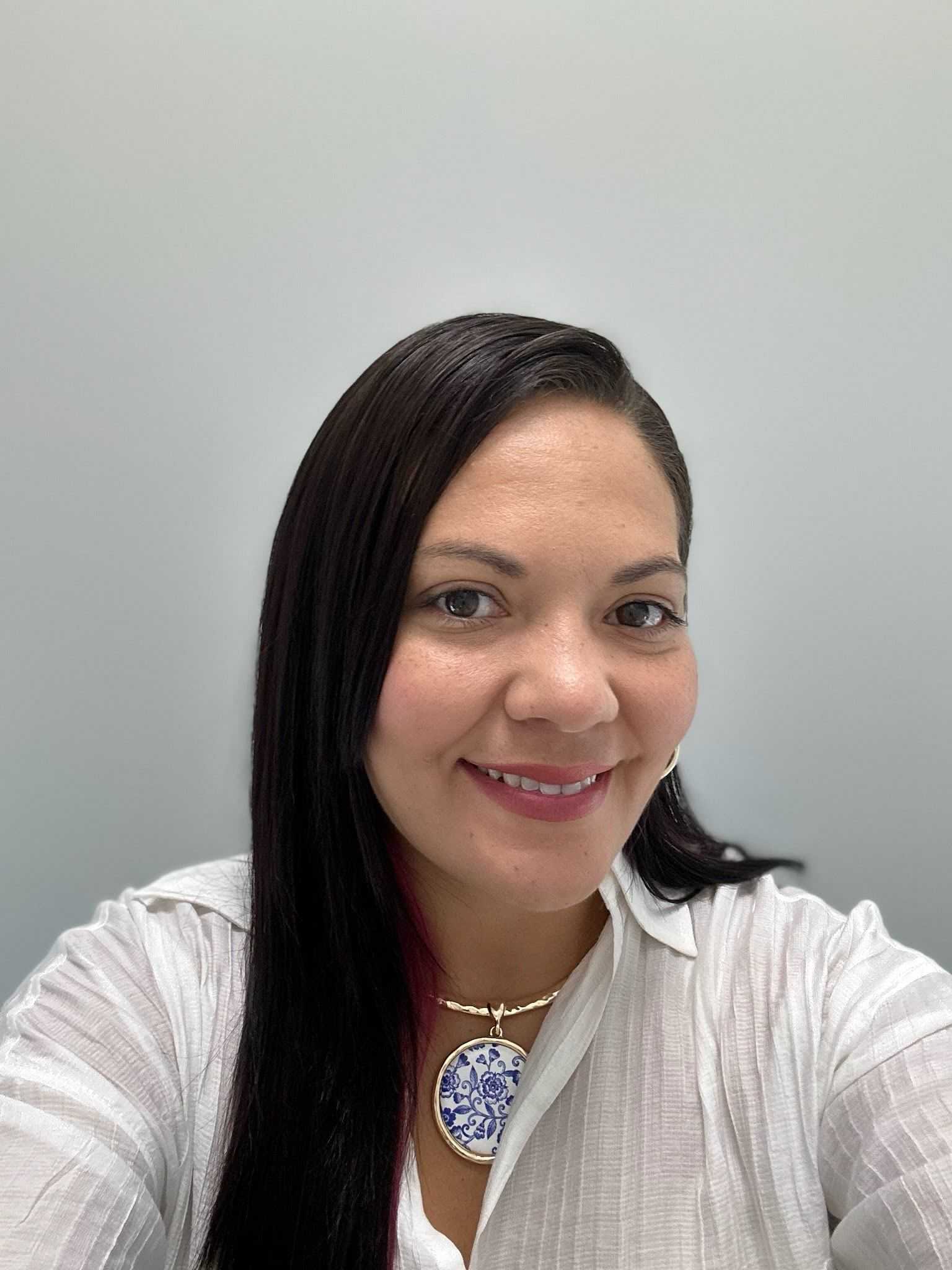 Woman smiling, wearing a white blouse and a blue pendant necklace.